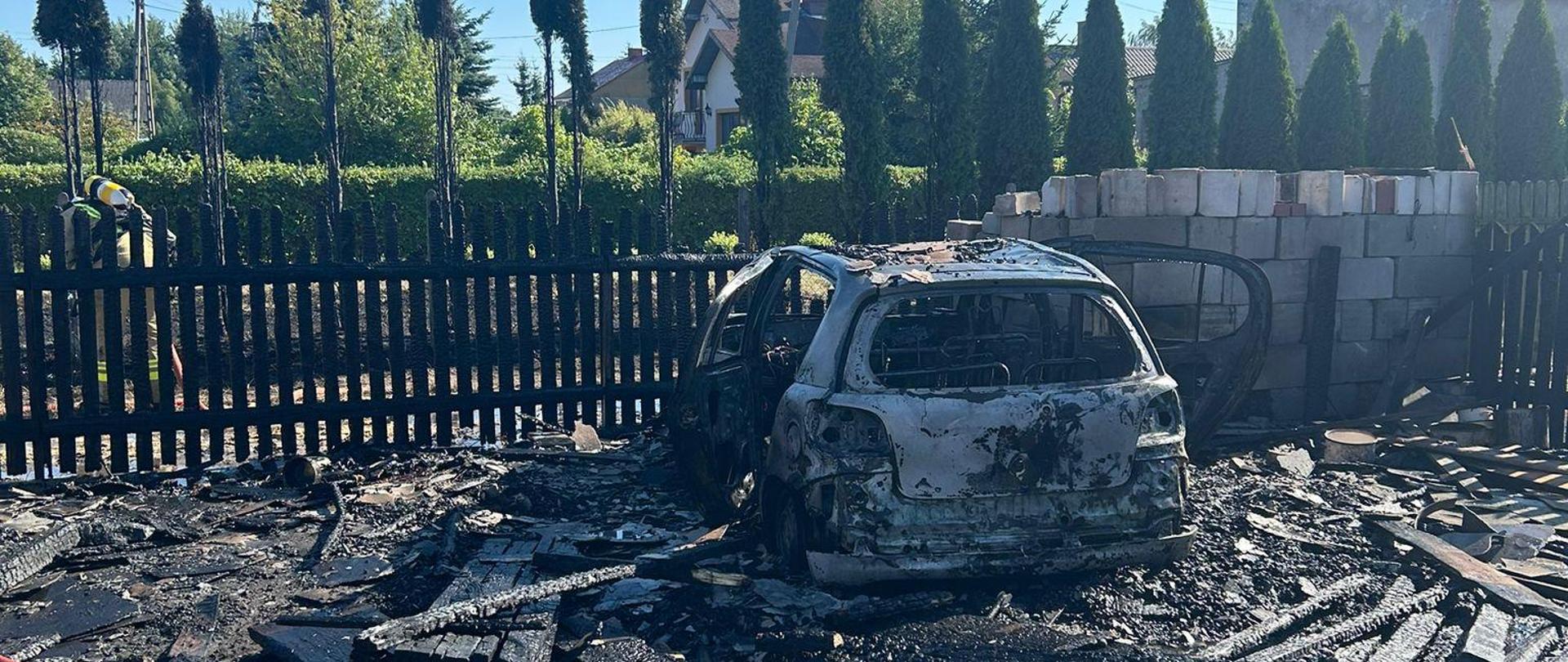 Słoneczna scena na świeżym powietrzu ukazuje zwęglone szczątki samochodu i budynku, prawdopodobnie garażu lub szopy, po pożarze. Szkielet samochodu, całkowicie spalony, leży pośród dużej sterty poczerniałego, roztrzaskanego drewna i gruzu. Ciemny drewniany płot, również noszący ślady spalenia, oddziela spalony obszar od rzędu zielonych drzew iglastych i budynków mieszkalnych w tle. Ziemia to mieszanka ziemi i popiołu. Po lewej stronie, częściowo widoczny jest strażak w piaskowym ubraniu specjalnym. Scena wyraźnie ukazuje niszczycielskie skutki pożaru.