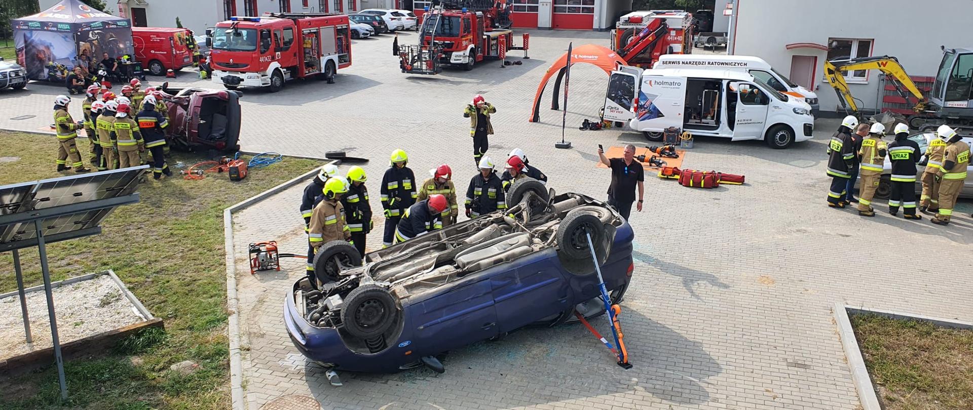 Zdjęcie przedstawia utwardzony plac, na którym odbywają się ćwiczenia strażackie. Przy wrakach 3 aut szkolą się ratownicy. W tle pojazdy strażackie, samochody osobowe i budynki strażnicy.