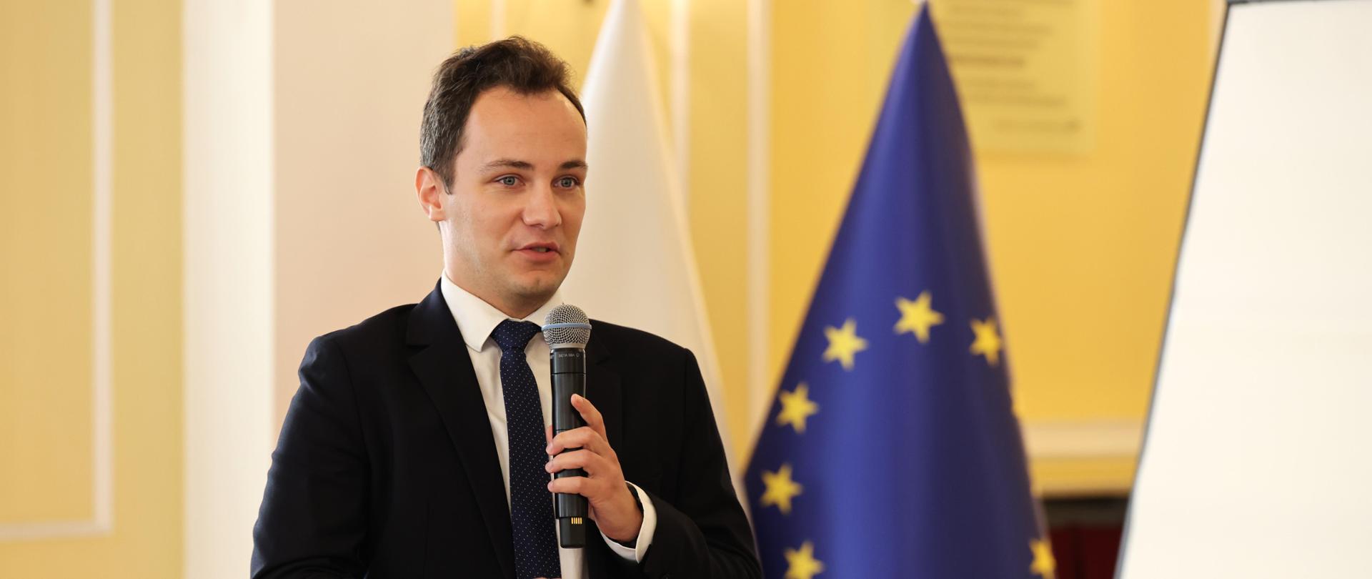 Wicewojewoda Patryk Fajdek przemawia do uczestników spotkania. w tle flaga Polski i Unii Europejskiej. 
