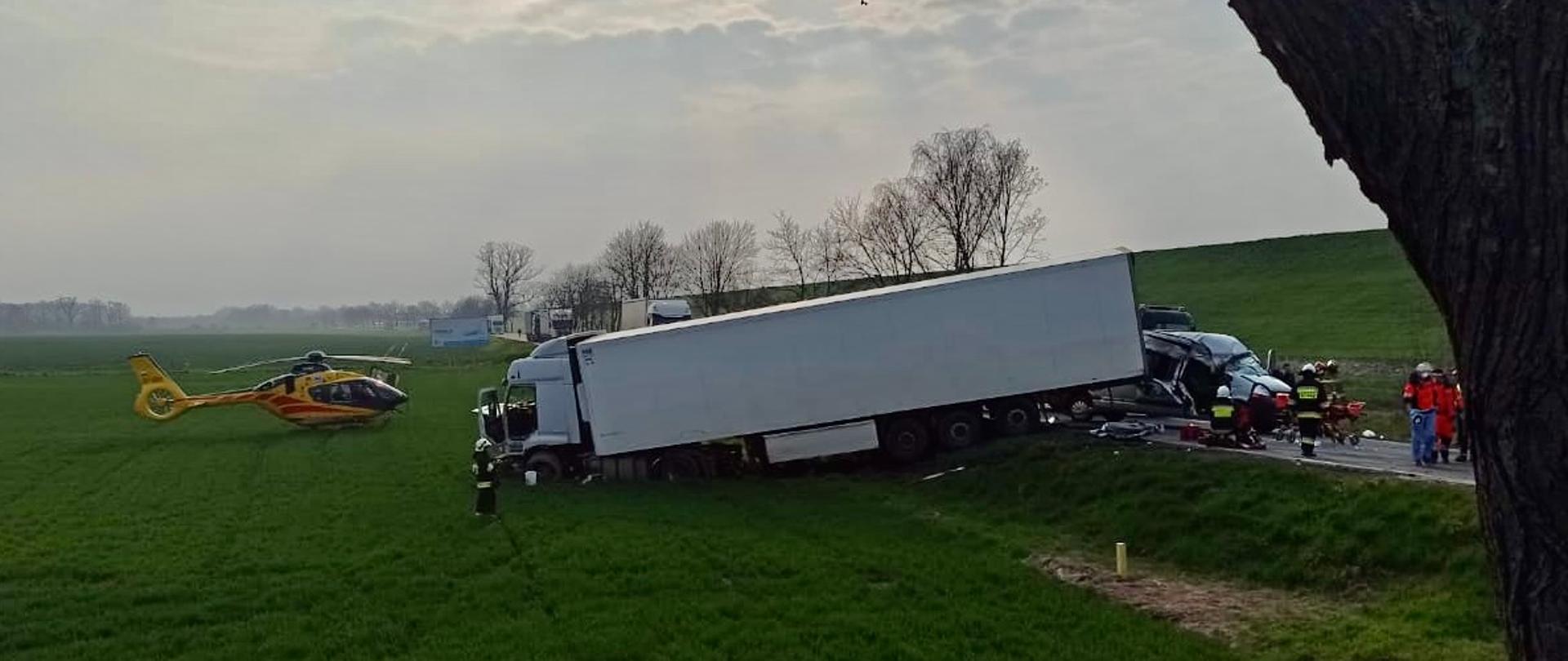 Zdjęcie przedstawia miejsce zdarzenia po wypadku samochodu ciężarowego, samochodu typu bus i samochodu osobowego. Po lewej stronie widoczny śmigłowiec Lotniczego Pogotowia Ratunkowego a po prawej widoczni strażacy i ratownicy medyczni.