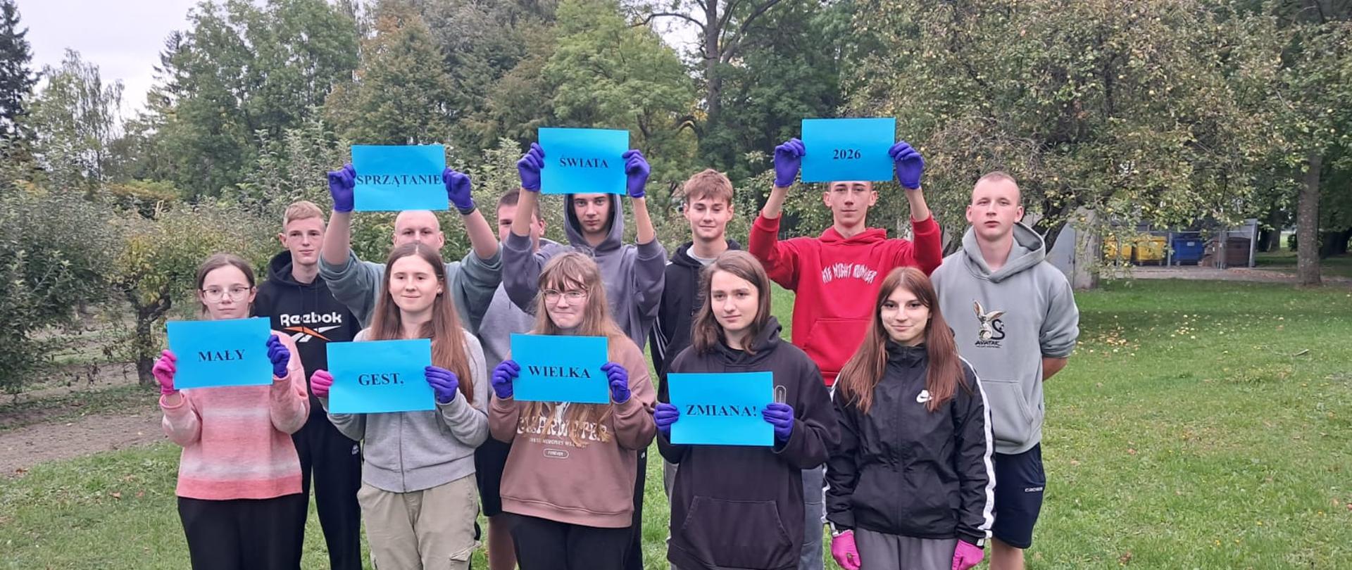 Grupa młodzieży pozująca na trawniku z niebieskimi kartkami tworzącymi hasło akcji ekologicznej.