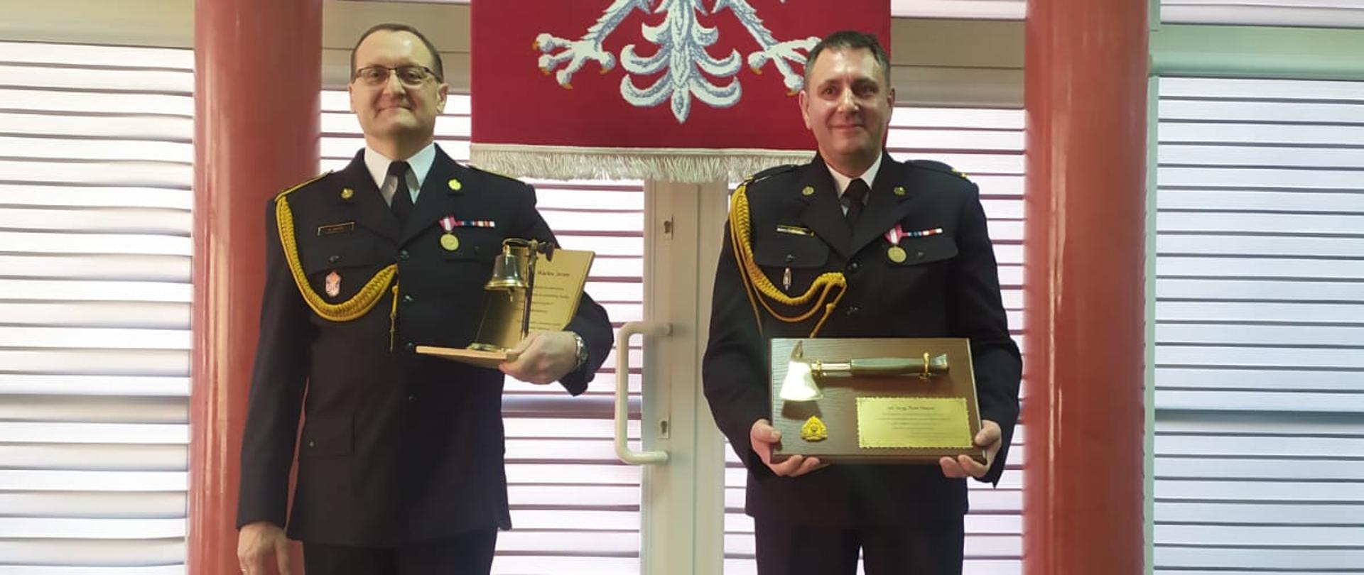 Na zdjęciu po prawej stronie stoi w umundurowaniu wyjściowym młodszy brygadier Piotr Mazur. W rękach trzyma pamiątkowy grawer z toporkiem strażackim. Po lewe stronie stoi w umundurowaniu wyjściowym aspirant sztabowy Wacław Jarosz. W lewej ręce trzyma pamiątkowy grawer z dzwonkiem. W tle za funkcjonariuszami Godło Polski oraz dwa czerwone filary.