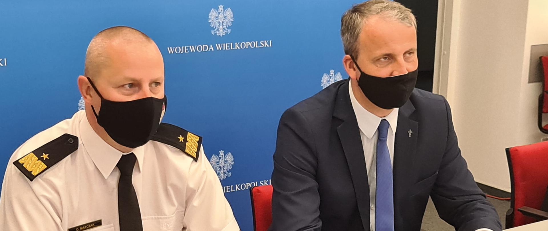 dwóch mężczyn z maseczkach ochronnych siedzi przy stole, jeden ubrany w białą koszulę z czarnym krawatem i dystynkcjami , drugi w garniturze z niebieskim krawatem