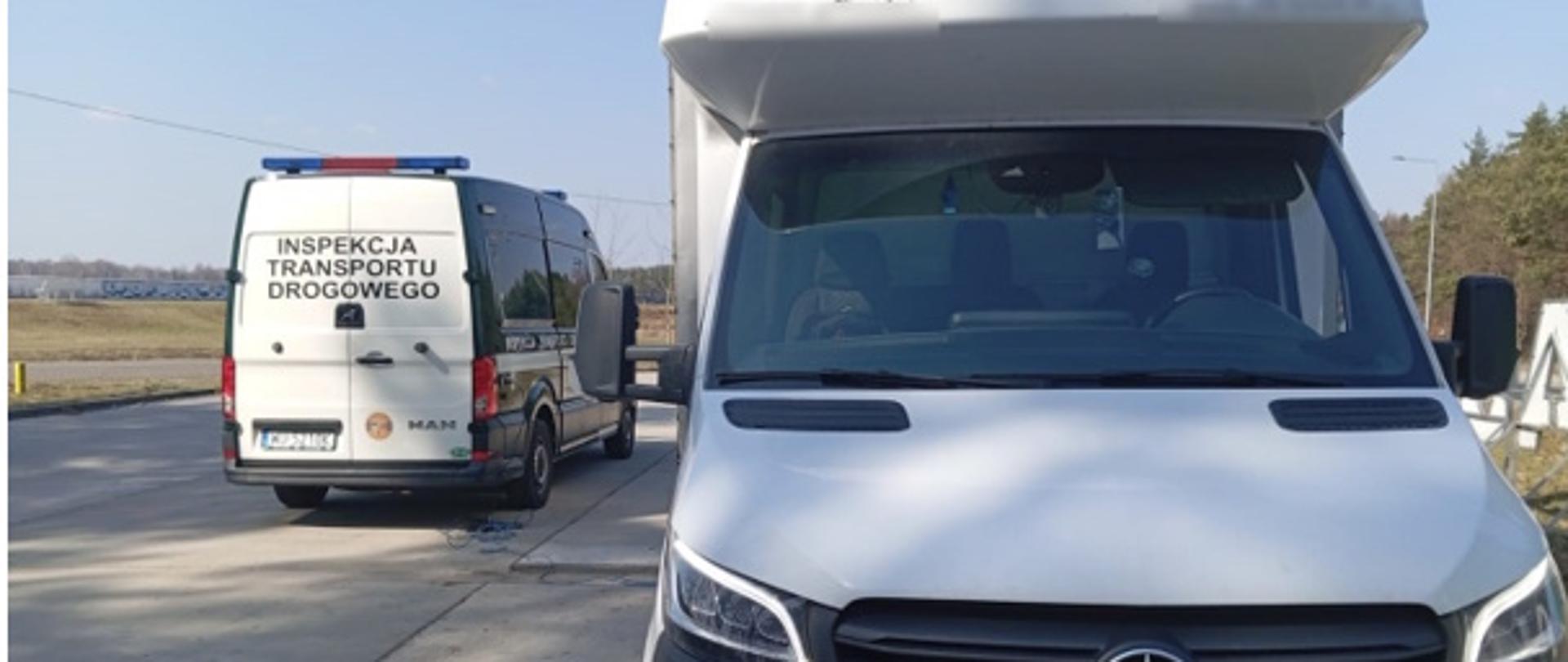 Na pierwszym planie znajduje się duży biały pojazd dostawczy do 3.5 t, ustawiony przodem do aparatu. Za nim, nieco dalej po lewej stronie, stoi pojazd z napisem „INSPEKCJA TRANSPORTU DROGOWEGO”.
Oba auta stoją na poboczu lub przy parkingu. W tle widać otwartą przestrzeń, prawdopodobnie pola, oraz kilka drzew po prawej stronie. 