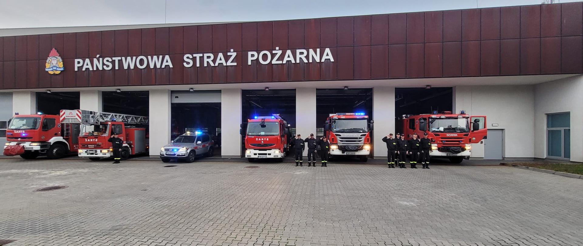 W tle budynek Komendy Miejskiej państwowej Straży Pożarnej, samochody strażackie w bramach z włączonymi światłami i strażacy oddający honor zmarłemu strażakowi. 