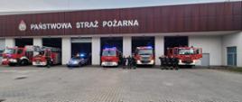 W tle budynek Komendy Miejskiej państwowej Straży Pożarnej, samochody strażackie w bramach z włączonymi światłami i strażacy oddający honor zmarłemu strażakowi. 