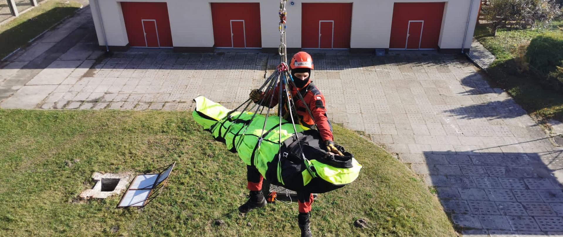 Ratownik wysokościowy ewakuuje na wysokości ( w powietrzu) na linie poszkodowanego (pozoranta) na noszach, w tle budynek biały z czerwonymi bramami