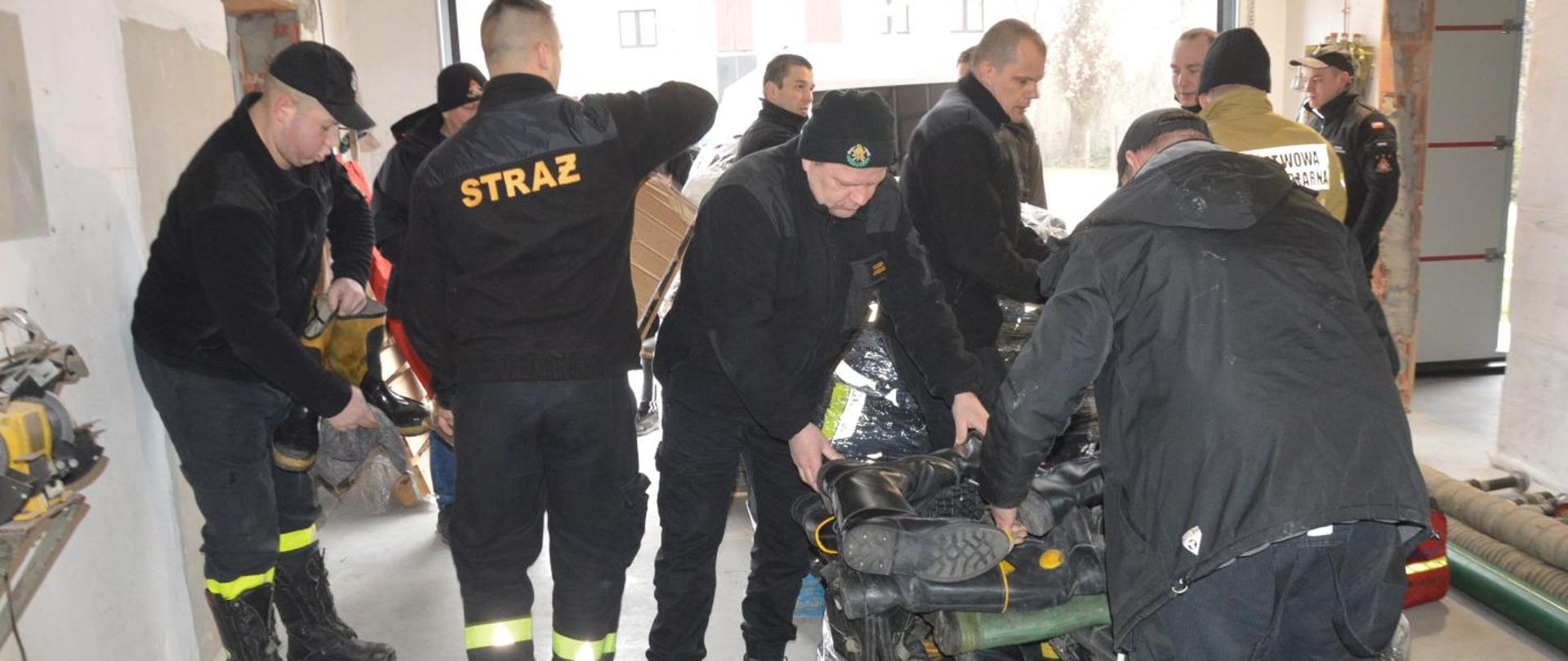 Sprzęt pożarniczy dla ukraińskich strażaków. Komenda Powiatowa PSP w Rawiczu. Strażacy przygotowują do transportu zebrany sprzęt. Na europalecie układają obuwie, które następnie będą owijać folią. W tle przyczepka i fragment budynku komendy.
