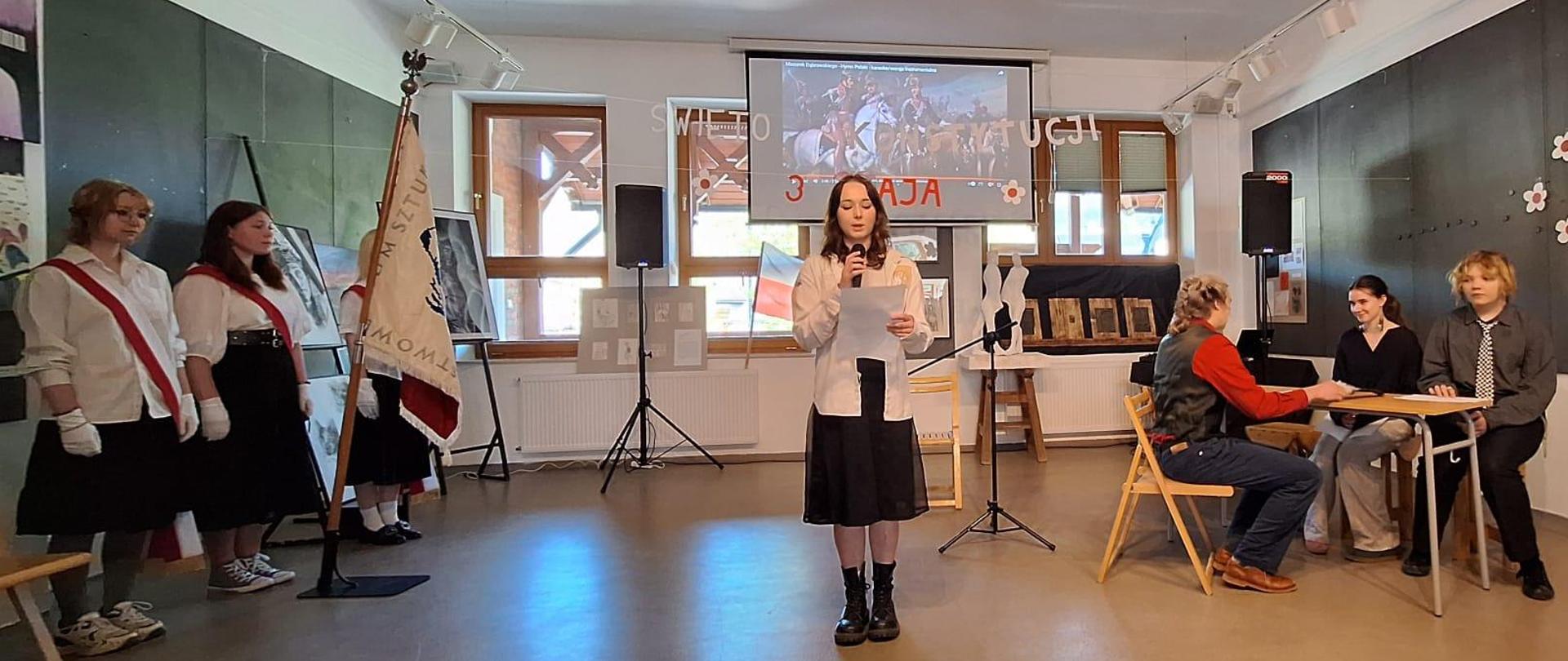 Na zdjęciu widzimy uczniów uczestniczących w szkolnej akademii z okazji Święta Konstytucji 3 Maja. Akademia odbywała się w szkolnej galerii "Strug" Po lewej stronie widzimy poczet sztandarowy składający się z trzech odświętnie ubranych uczennic. Uczennice ubrane są w białe koszule i granatowe spódnice, są również przepasane przez ramię biało-czerwonymi szarfami. Na rękach maja założone białe rękawiczki. Przed uczennicami na stojaku stoi szkolny sztandar. Na środku widzimy uczennicę odświętnie ubraną i trzymającą w ręku mikrofon, która czyta tekst związany z uroczystością 3 Maja. Po prawej stronie przy stoliku siedzą uczniowie. W tle widzimy litery zawieszone na sznurku. Litery tworzą napis biało-czerwony: Święto Konstytucji 3 Maja, dalej widzimy również ekran z wyświetlanym filmem dotyczącym uchwalenia Konstytucji 3 Maja 