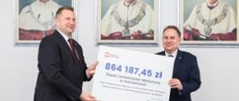 Minister Czarnek i mężczyzna w garniturze stoją i trzymają razem symboliczny czek z napisem 864 187,45 zł, za nimi na ścianie wiszą 3 portrety.