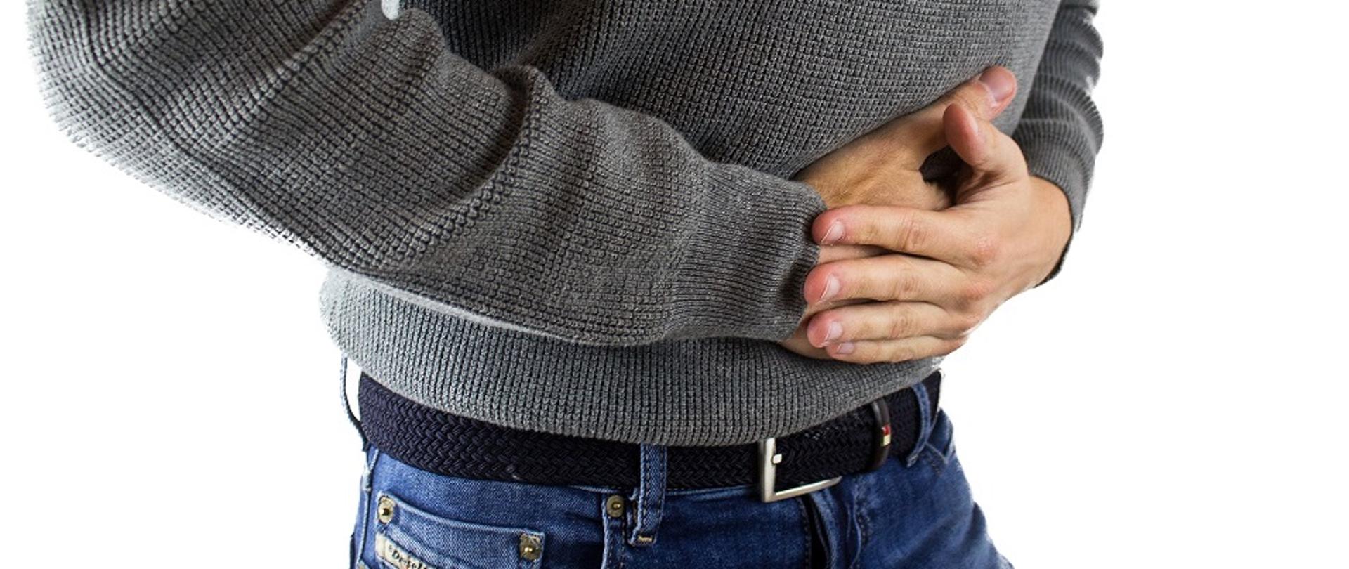 fragment postaci mężczyzny, lekko zgiętej w pasie.
Mężczyzna trzyma się obiema rękoma za brzuch.
