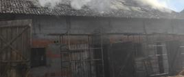 Zdjęcie przedstawia pożar budynku inwentarskiego. Nad dachem budynku widać zadymienie. Przed budynkiem stoi rusztowanie. 