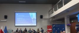 Duża grupa ludzi siedzi w sali, zwrócona w stronę prelegentów. 