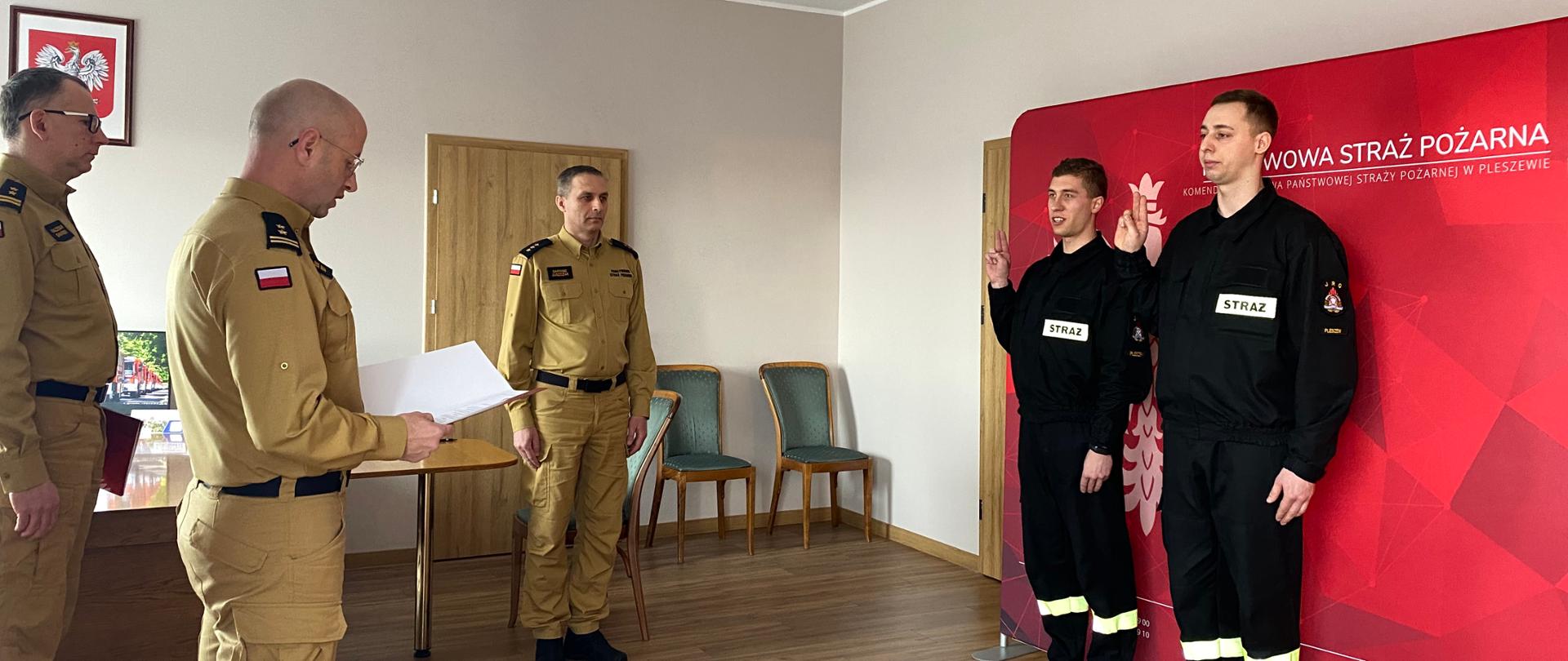 Dzień, pomieszczenie biurowe, dwóch strażaków ubranych w czarne mundury koszarowe stoi na tle czerwonego baneru, trzymając prawe ręce w geście ślubowania. Przed nimi stoi trzech mężczyzn w mundurach straży pożarnej koloru musztardowego, jeden z mężczyn odczytuje trzymany w ręku dokument