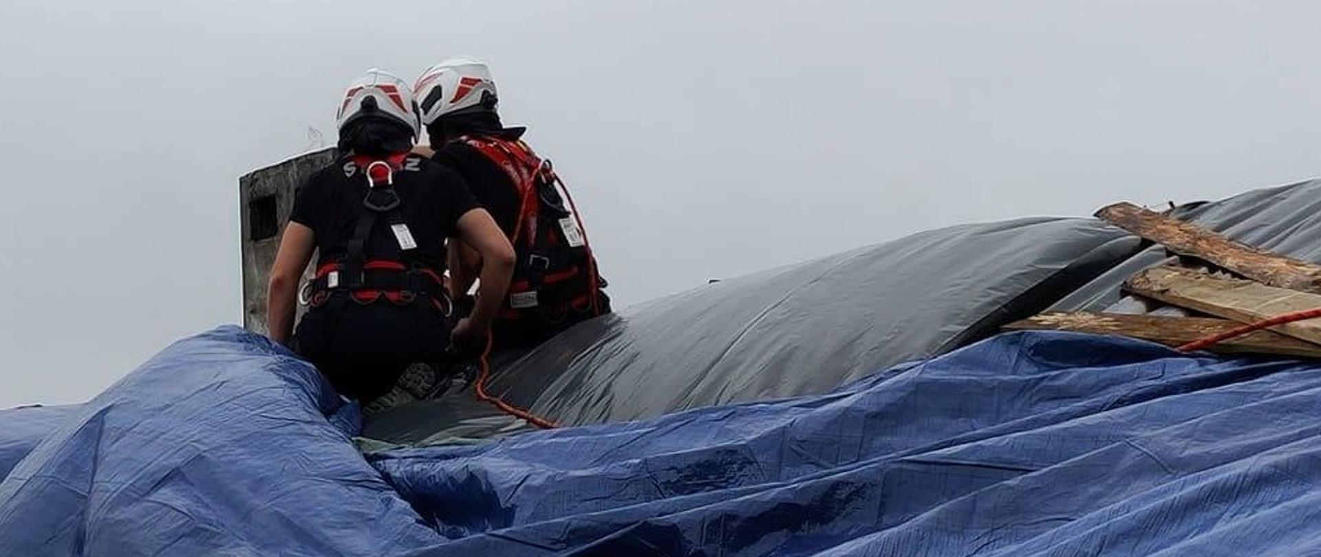 Zdjęcie przedstawia dwóch strażaków ubranych w sprzęt ratownictwa wysokościowego znajdujących się na uszkodzonym dachu budynku i zabezpieczających poszycie plandekami