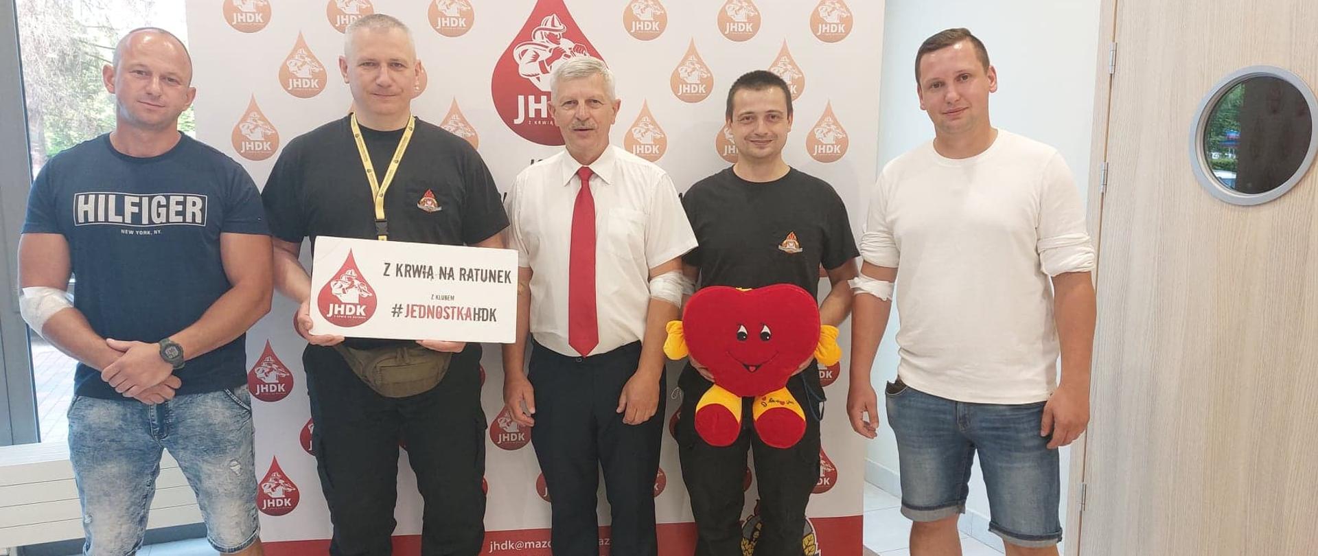 Grodziscy strażacy wraz z prezesem Klubu Jednostka HDK stoją na tle ścianki z kroplami krwi w których jest wizerunek strażaka i napis JHDK. Jedna osoba trzyma białą tabliczkę z napisem "z krwią na ratunek z Klubem #JEDNOSTKAHDK", druga osoba trzyma maskotkę w kształcie kropli krwi. 