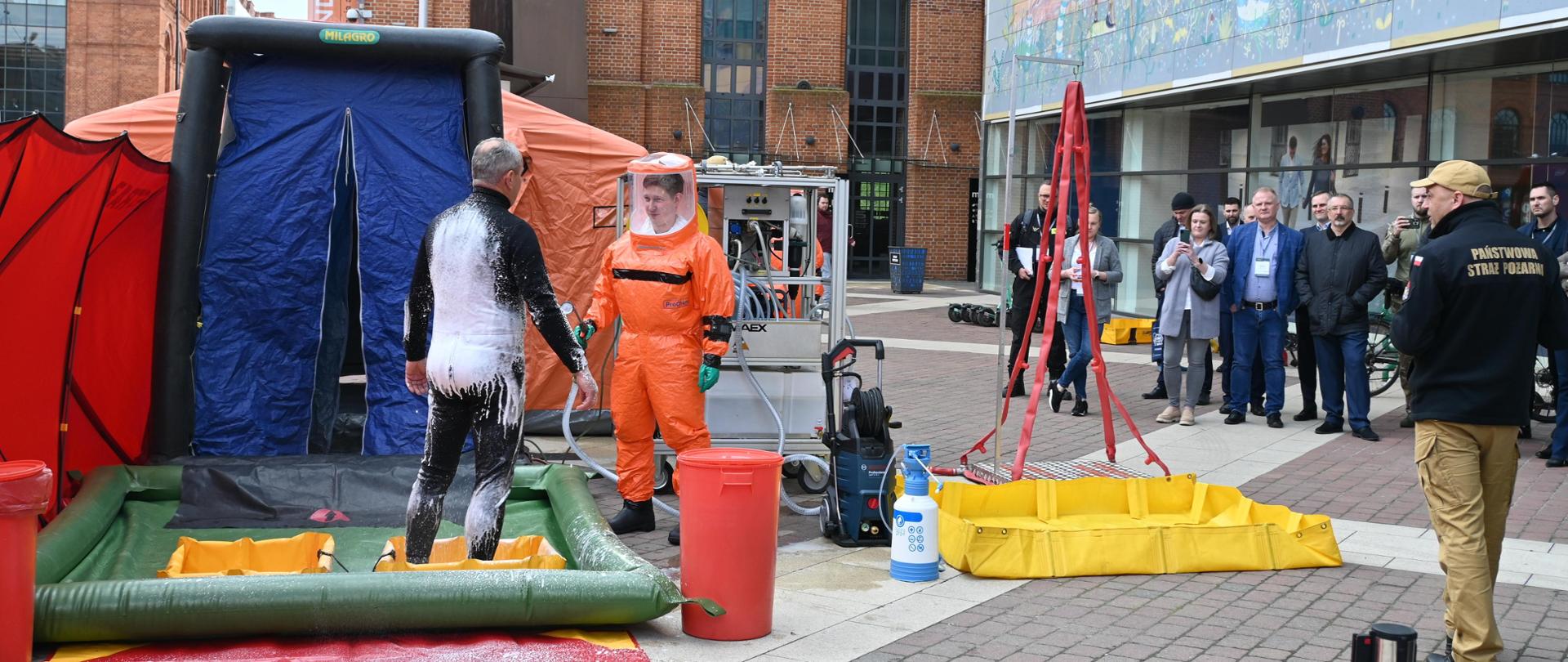 Strażak prowadzący dekontaminację mężczyzny przed namiotem, po prawej stronie osoby obserwujące ćwiczenia