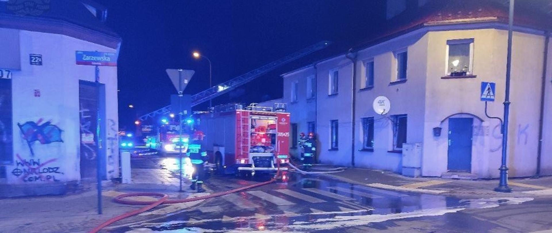 Samochód ratowniczo-gaśniczy stojący na ulicy pomiędzy budynkami, samochód podłączony wężem do hydrantu, obok samochodu strażacy. W tle wysunięta drabina w kierunku jednego z budynków