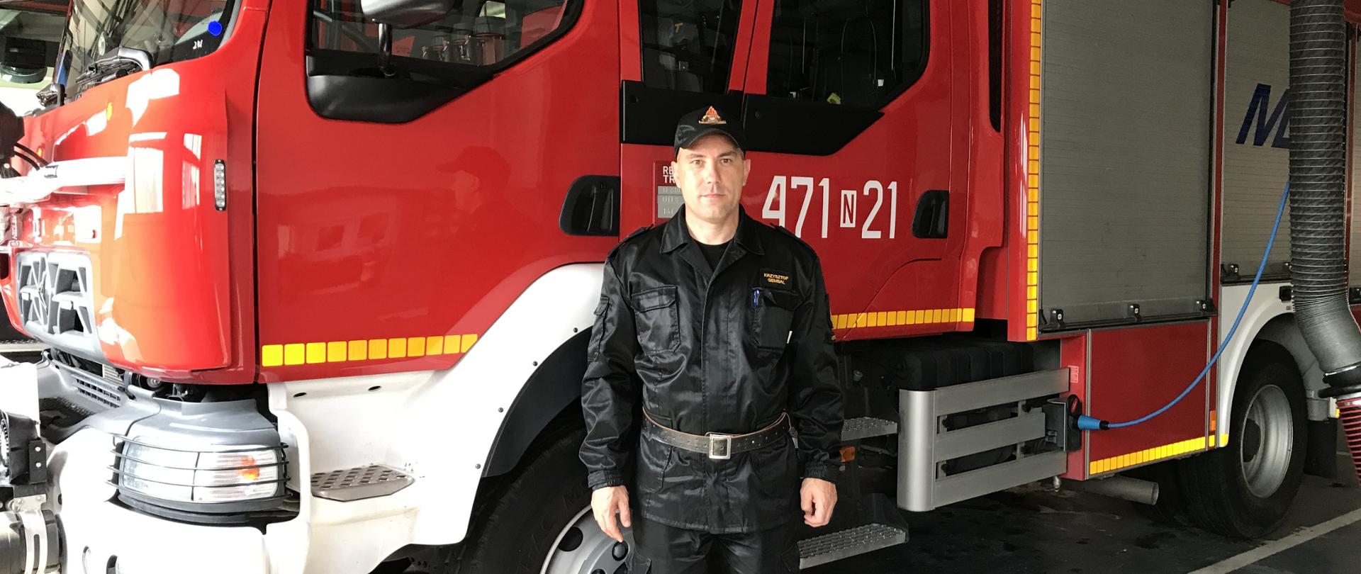 Strażak ubrany w czarne ubranie dowódczo-sztabowe na tle czerwonego wozu ratowniczo-gaśniczego.