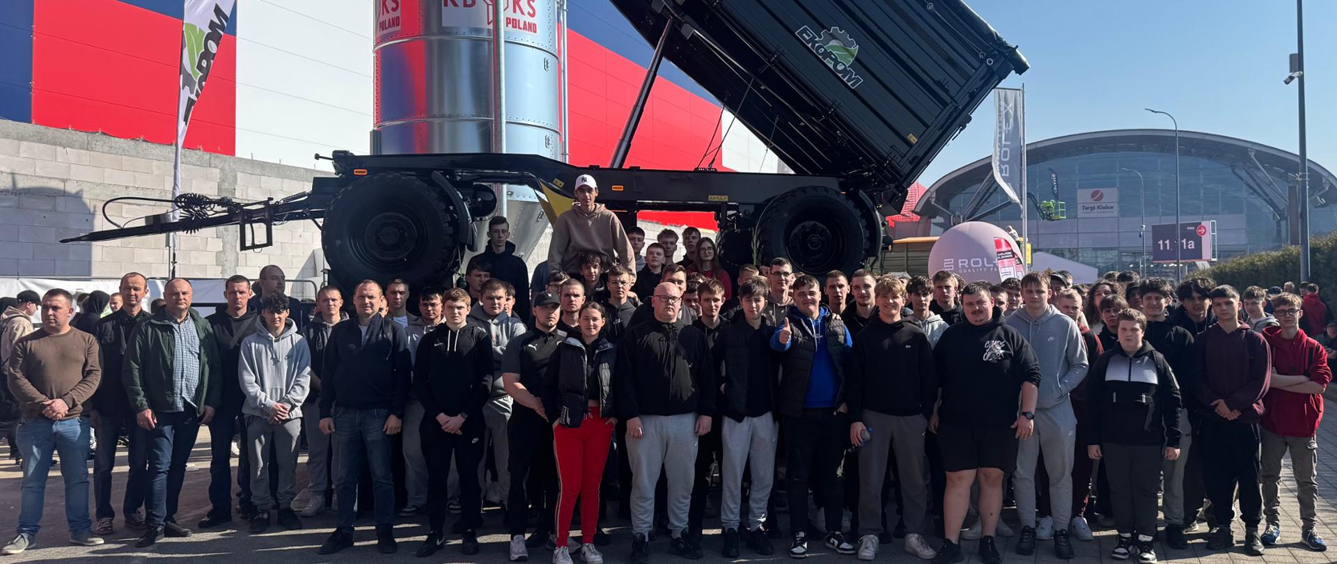 Duża grupa osób stoi przed maszyną rolniczą z uniesioną skrzynią, ustawioną na zewnątrz hali targowej.