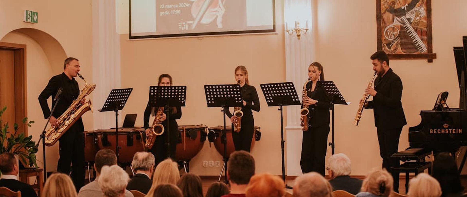 Zespół pięciu muzyków grających na scenie auli na saksofonach, w tle na ciemnożółtej ścianie obraz o tematyce muzycznej i a na ekranie widoczny plakat z wydarzenia, pod sceną auli widoczna publiczność.