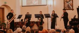 Zespół pięciu muzyków grających na scenie auli na saksofonach, w tle na ciemnożółtej ścianie obraz o tematyce muzycznej i a na ekranie widoczny plakat z wydarzenia, pod sceną auli widoczna publiczność.
