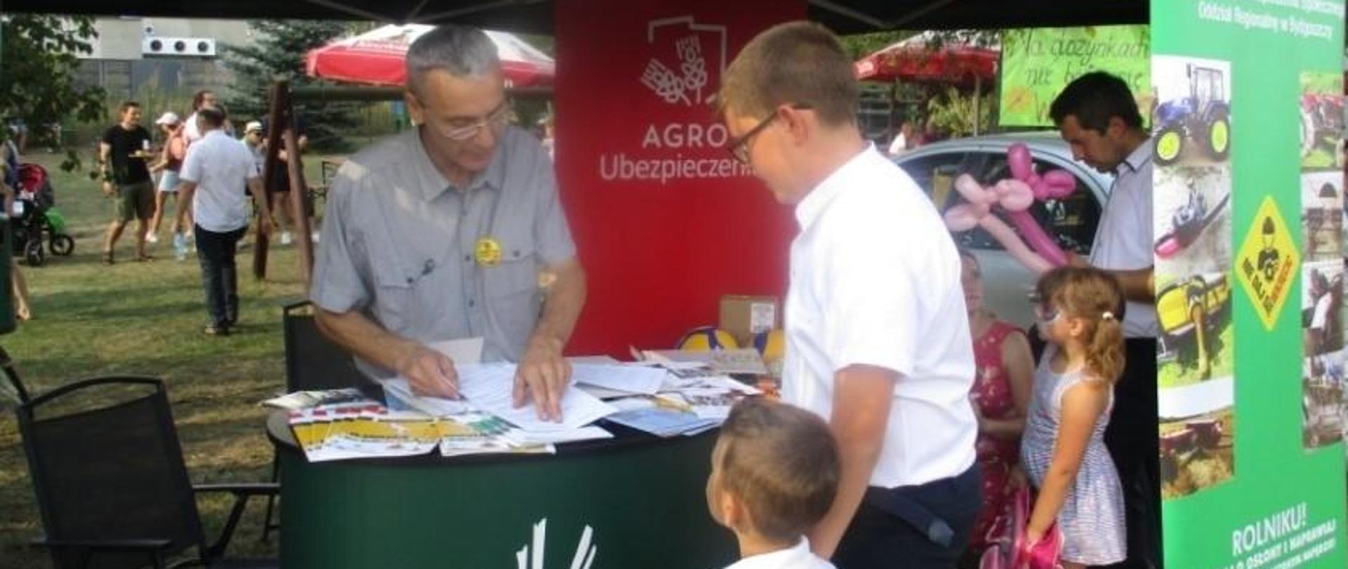 W namiocie z logo KRUS pracownik Placówki rozmawia z dwójką dzieci. Na blacie stolika wyłożone są ulotki prewencyjne.