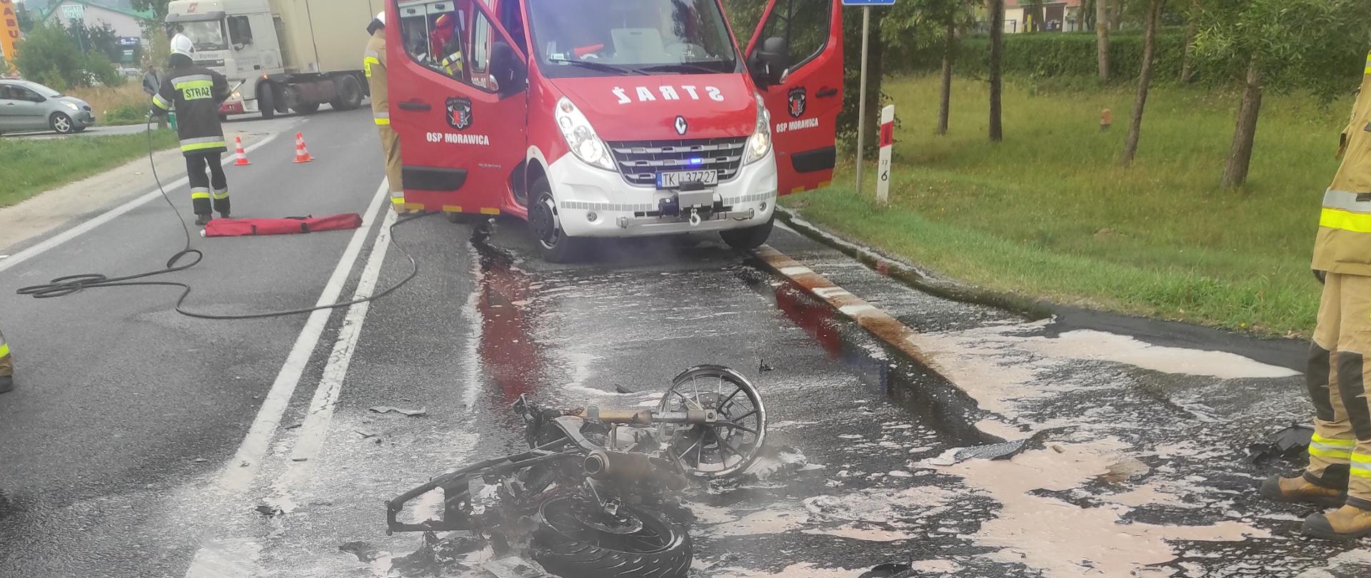 Zdjęcie przedstawia spalony motocykl na jezdni. Za nim wóz strażacki.