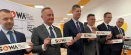 Sześciu uśmiechniętych mężczyzn w garniturach stoi w rzędzie, każdy trzyma kawałek taśmy z napisem SOWA, za nimi duż baner z takim samym napisem.