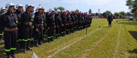Zdjęcie przedstawia druhów OSP ustawionych w trzyszeregu. Ubrani są w ubranie koszarowe, pasy strażackie oraz hełmy. Strażacy stoją na boisku z trawą.