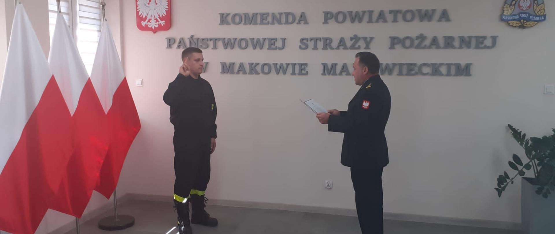 Strażak podnosi prawą rękę z wyciągniętymi dwoma palcami w postawie zasadniczej. Komendant Powiatowy z dokumentem w teczce trzymanym w rękach stoi na wprost niego. W tle napis na ścianie "Komenda Powiatowa Państwowej Straży Pożarnej w Makowie Mazowieckim" oraz godło Polski i logo PSP.