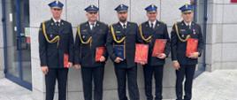 Na zdjęciu strażacy w galowych mundurach. Strażacy w rękach trzymają dyplomy. W tle marmurowa, świecąca się ściana szkoły z dużą nazwą jednostki. 