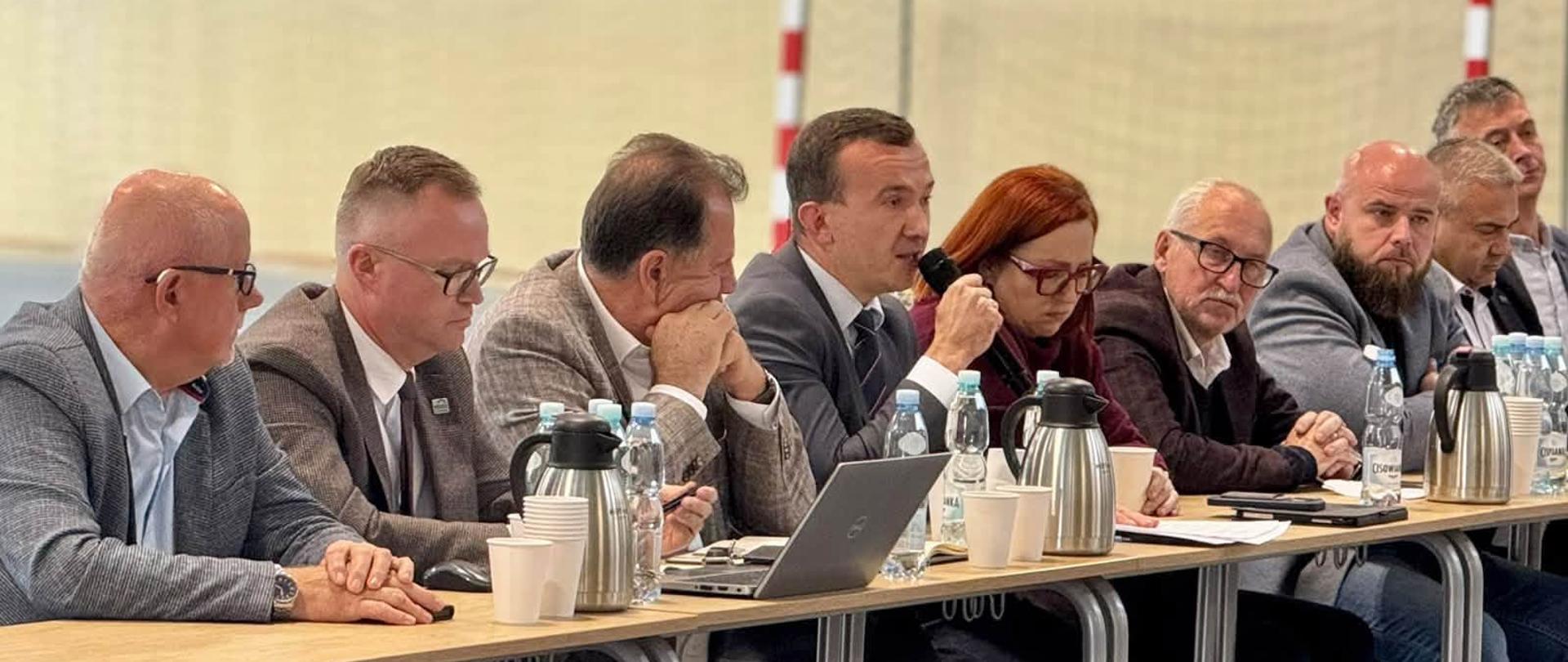 grupa oficjeli, w tym wicewojewoda, siedzą przy stole, wicewojewoda przemawia przez mikrofon