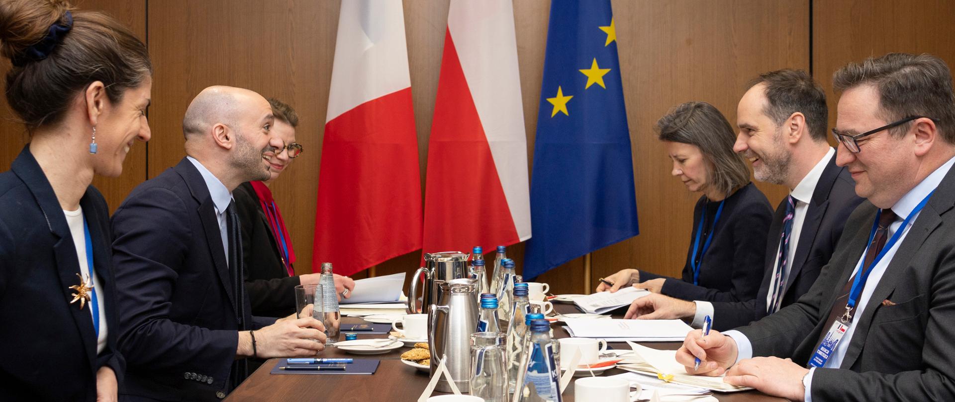 Delegacje FR i PL siedzą przy stole konferencyjnym podczas spotkania, rozmawiając w obecności flag Francji, Polski i Unii Europejskiej. Na zdjęciu widać Ministra Delegowanego do Spraw Europy Republiki Francuskiej Benjamina Haddada z dwiema kobietami siedzącymi po jego obu stronach oraz Sekretarza Stanu w Ministerstwie Spraw Zagranicznych Ignacego Niemczyckiego obok którego siedzi mężczyzna i kobieta.