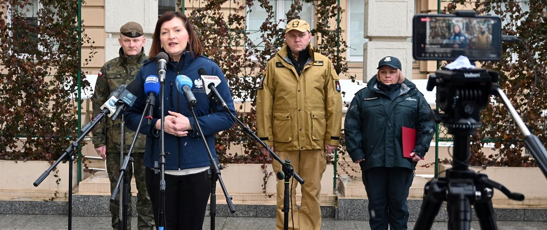 Zdjęcie zrobione na zewnątrz. Na pierwszym planie czworo uczestników konferencji prasowej. Wśród nich oficerowie: Państwowej Straży Pożarnej, Wojsk Obrony Terytorialnej i Izby Skarbowej ubrani w mundury. Przed mikrofonami stoi wojewoda podkarpacki.