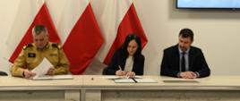 Strażak kobieta mężczyzna siedzą przy stole podpisują dokumenty za nimi znajdują się flagi Polski oraz część ekranu z logiem Państwowej Straży Pożarnej.