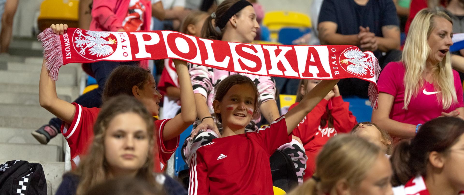 Na stadionowej trybunie siedzą dzieci, dwóch chłopców w czerwonych koszulkach trzyma rozpostarty biało-czerwony szalik z napisem Polska.
