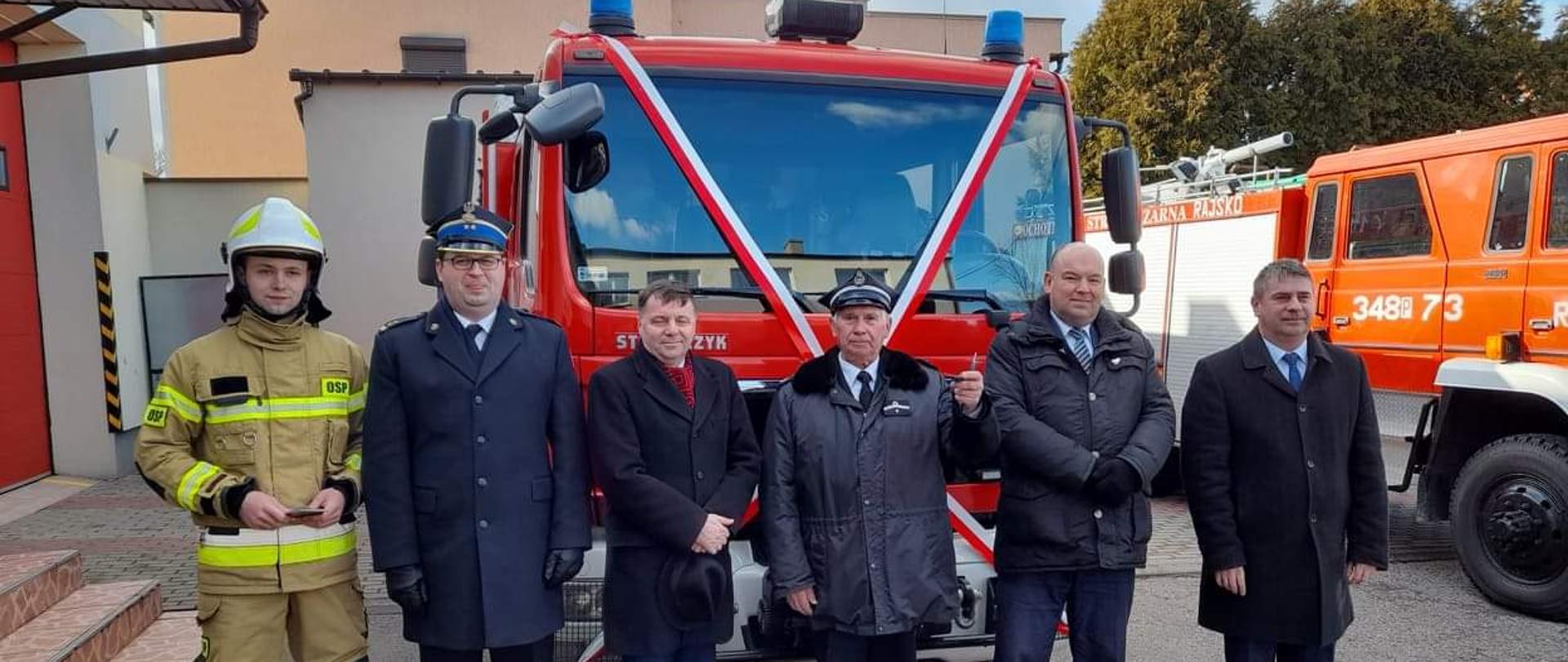 przed czerwonym samochodem z biało-czerwona szarfą stoi grupa mężczyzn, wśród nich strażak w jasnym umudurowaniu bojowym i hełmnie, strażak w rogatywce i w plasczu