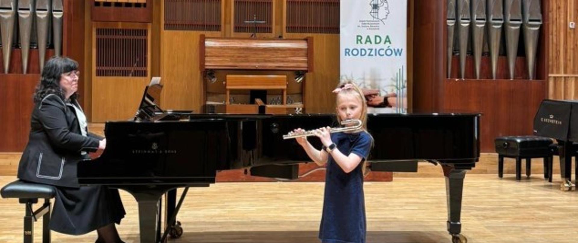 Zdjęcie kolorowe. Dziewczynka z długimi, blond włosami, ubranymi w ciemną sukienkę, stoi na scenie i gra na flecie poprzecznym. Obok niej siedzi kobieta, ubrana w czarny garnitur, grająca na czarnym fortepianie. Za nimi, na ścianie z jasnego drewna, organy z metalowymi piszczałkami. Po prawej stronie, za fortepianem, widoczny jest czarny baner z napisem „RADA RODZICÓW”.
