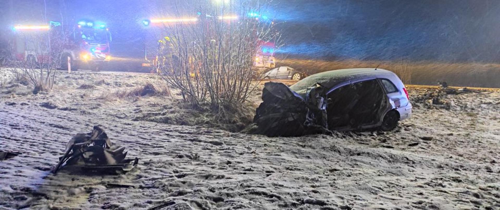 Na zaśnieżonym poboczu drogi leży rozbity samochód osobowy. Z tyłu stoją dwa czerwone wozy bojowe z włączonymi sygnałami świetlnymi.