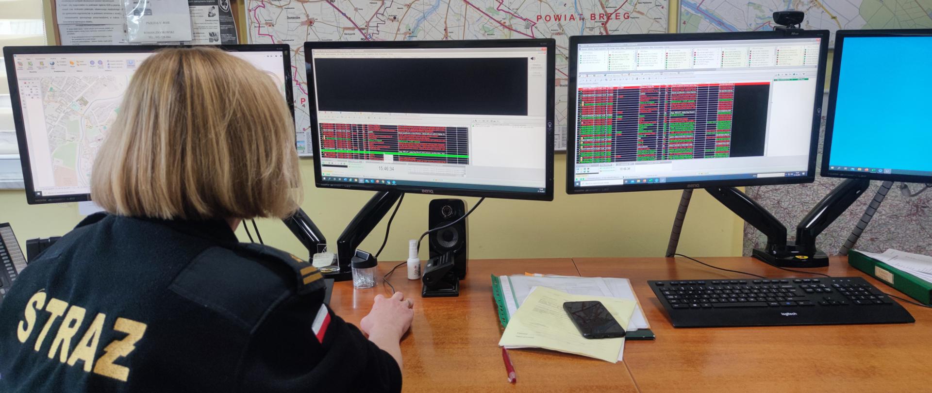 na zdjęciu widać Dyżurną straży pożarnej na stanowisku kierowania. Widać cztery monitory, na ekranach system ewidencji zdarzeń i dysponowania.