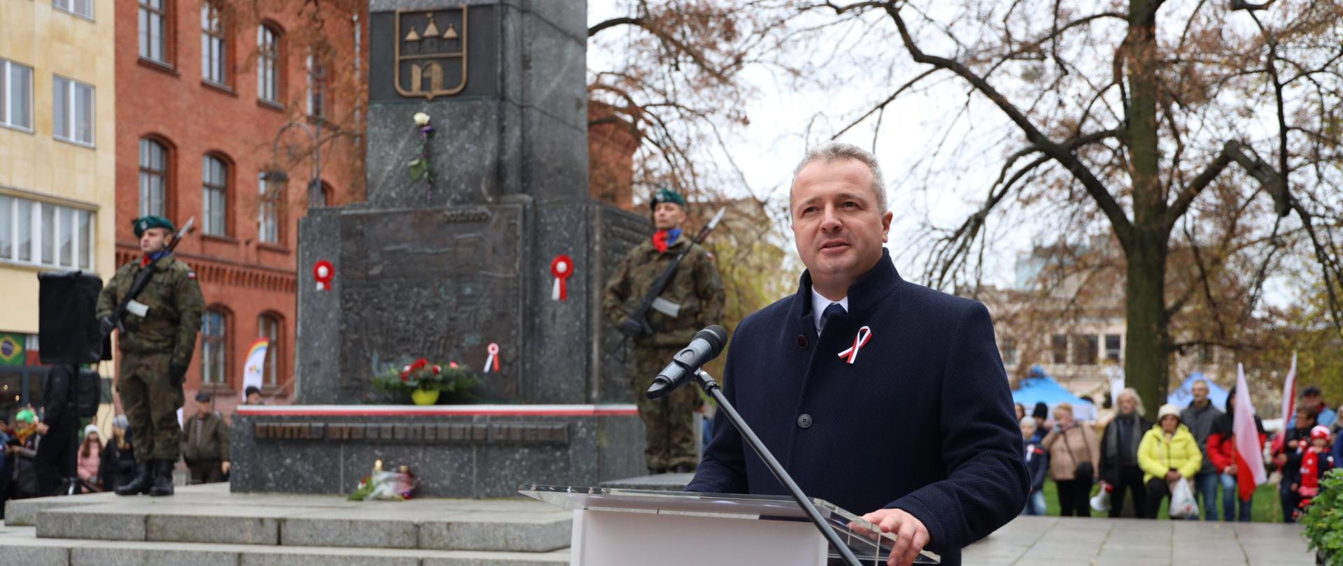 Jeden mężczyzna stojący przy mównicy i wypowiadający się. Obok wiele osób