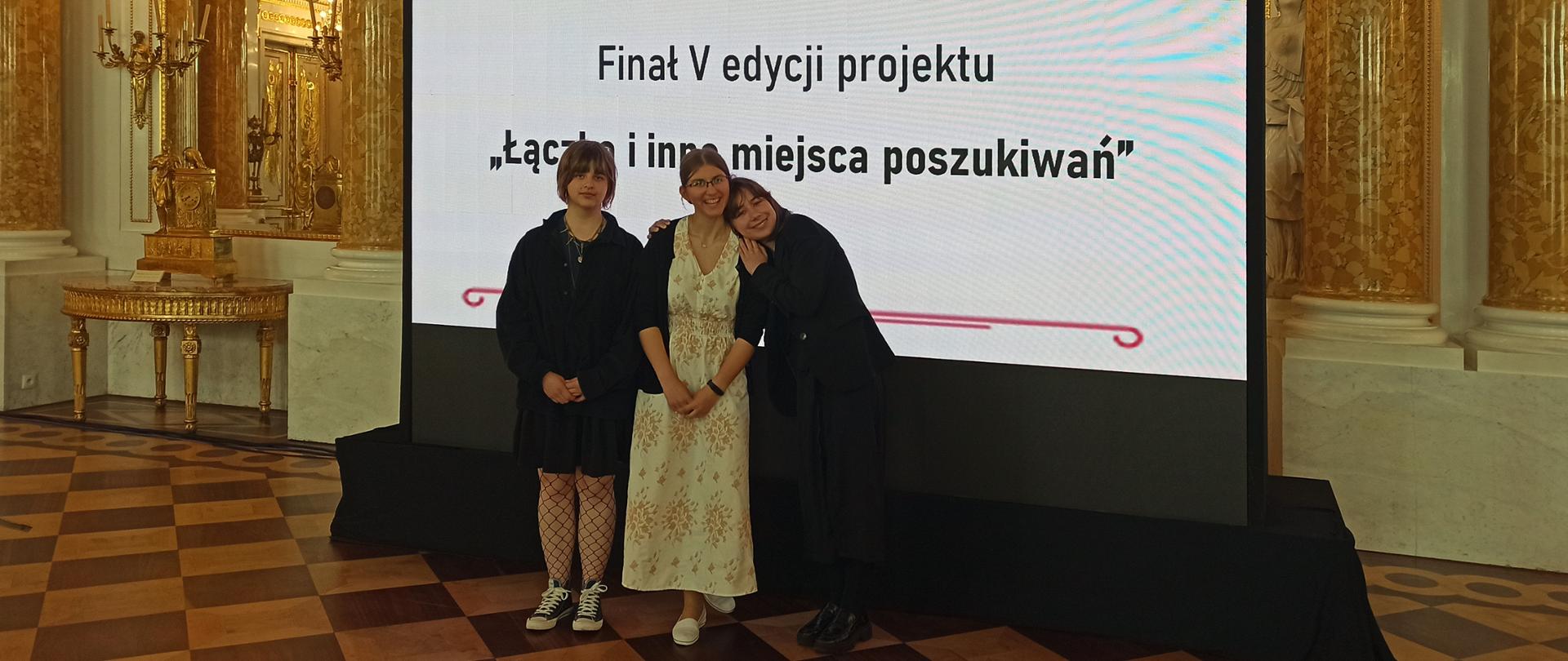 Trzy uczennice w Sali Wielkiej Zamku Królewskiego w Warszawie na tle telebimu z napisem Finał V edycji projektu "Łączka i inne miejsca poszukiwań".