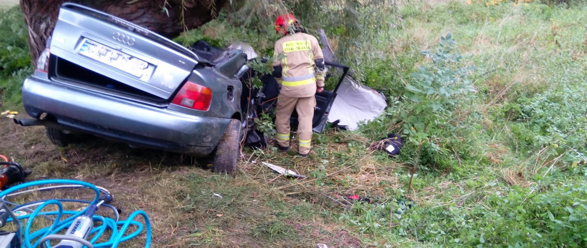 Srebrne auto z rozbitą prawą stroną, obok umundurowany strażak