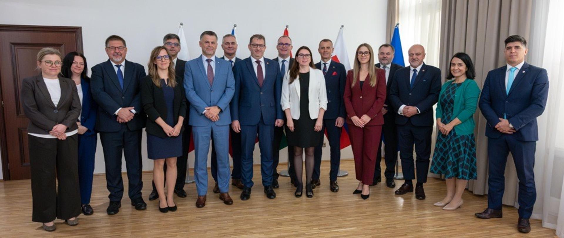 Grupa uczestników stoi w sali konferencyjnej do wspólnego zdjęcia. W tle widoczne są flagi kilku państw ustawione przy ścianie.