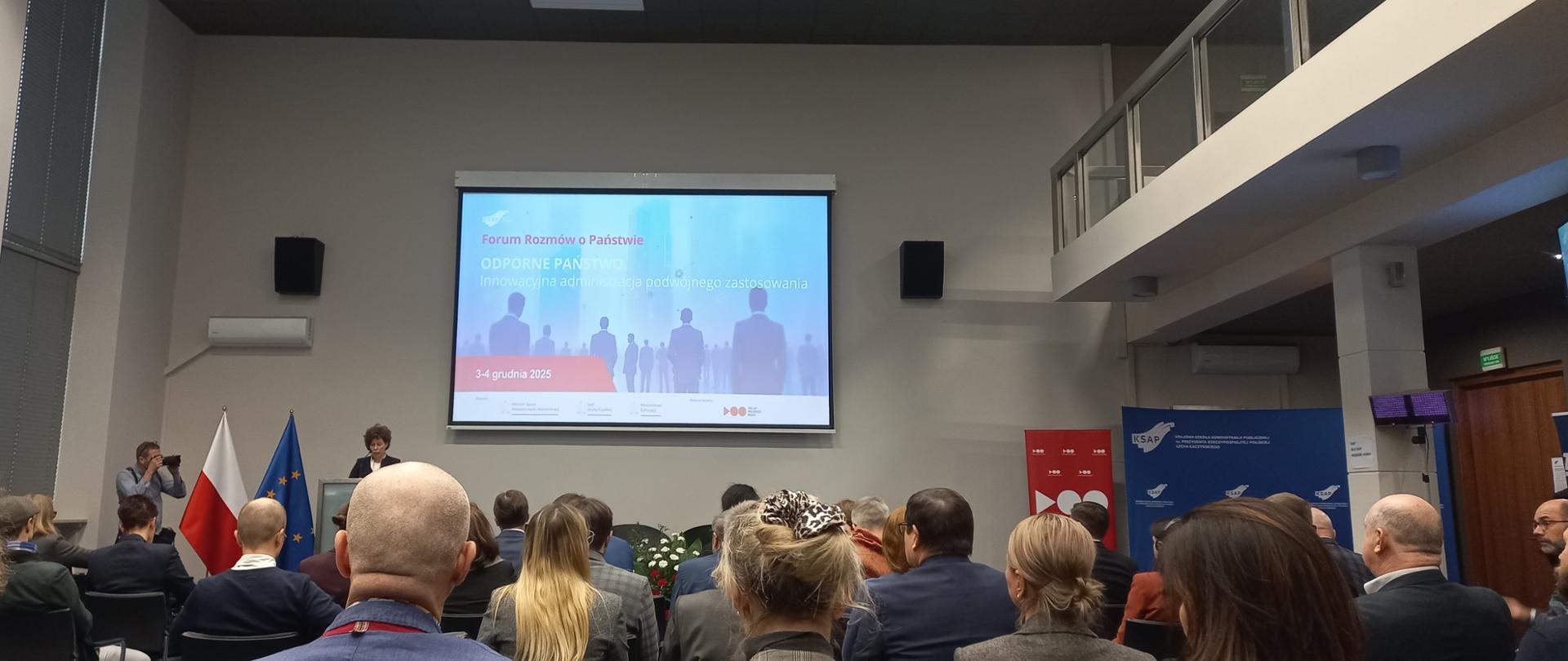 Grupa ludzi siedzi w pomieszczeniu, uczestnicząc w spotkaniu lub wykładzie. 