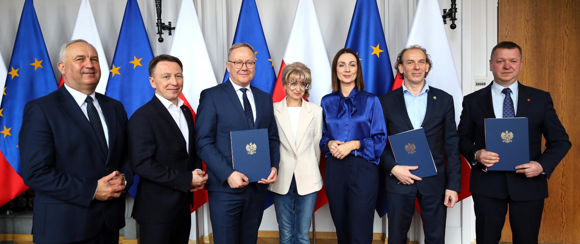 kilka osób stoi na tle flag i trzyma w dłoniach podpisane umowy o dofinansowaniu inwestycji