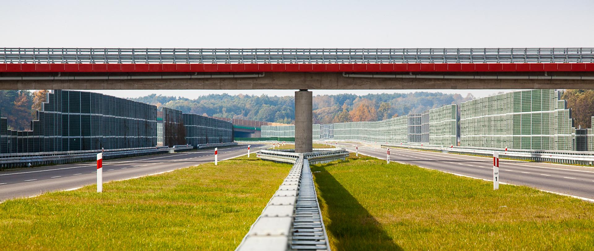 Widok z poziomu ziemi na autostradę. Zdjęcie zrobione z pasa rozdziału między jezdniami na którym rośnie trawa. Po środku jest bariera energochłonna. W dalszej perspektywie widać wiadukt nad autostradą. Po obu stronach autostrady są zlokalizowane ekrany akustyczne. w Tle widać kolorowe korony drzew co oznacza, że jest początek jesieni. Świeci słońce. 