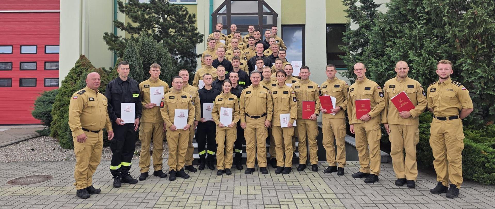 Grupa 39 strażaków w mundurach służbowych stoi przed budynkiem. Część z nich trzyma w ręku białe teczki. W centralnym miejscu stoi Komendant Szkoły.
