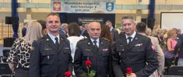 Trzech mężczyzn w galowych mundurach straży pożarnej pozuje do zdjęcia wewnątrz sali gimnastycznej podczas uroczystości inauguracji roku szkolnego 2025/2026. Każdy z nich trzyma w ręce czerwoną różę. W tle widać tłum osób w eleganckich strojach rozmawiających i przemieszczających się między rzędami krzeseł. Na ścianie wisi baner z napisem „Wojewódzka Inauguracja Roku Szkolnego 2025/2026” oraz herbami województwa i powiatu. Atmosfera wydarzenia jest oficjalna i uroczysta.